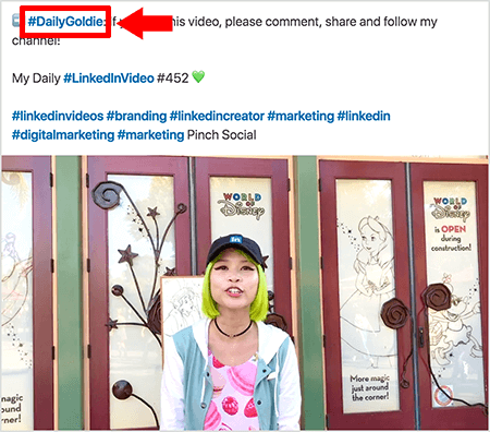 Detta är en skärmdump som illustrerar hur Goldie Chan använder hashtags i texten i sina LinkedIn-videoposter. Röda bildtexter pekar på #DailyGoldie-hashtaggen i texten, vilket är unikt för hennes videoposter och hjälper henne att spåra aktier. Inlägget innehåller också andra relevanta hashtags som hjälper människor att hitta hennes video, inklusive #LinkedInVideo. I videobilden står Goldie framför några dörrar vid en World of Disney-skärm. Hon är en asiatisk kvinna med grönt hår. Hon har en svart LinkedIn-keps, ett svart chokerhalsband, en rosa skjorta med makarontryck och en blå och vit jacka.
