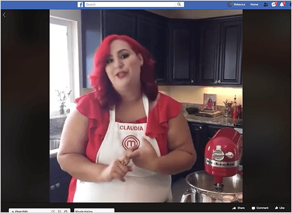 Detta är en skärmdump av en Facebook Live-video med kocken Claudia Sandoval, som hon var värd 2016 som en del av en tvärkampanj med T-Mobile Tirday-appen. I videon står Claudia i ett kök med svarta skåp och bänkskivor i granit. Ett fönster över diskbänken släpper in naturligt ljus i rummet. Claudia står bredvid en röd Kitchen Aid-mixer. Hon är en latinsk kvinna som har ljusrött hår som faller strax under hennes axlar. Hon har smink, en ljusröd topp och ett vitt förkläde med Claudia och MasterChef-logotypen broderad i röd tråd. När hon pratar gester hon med händerna. År 2016 samarbetade Stephanie Liu med Claudia för att producera denna livevideo.
